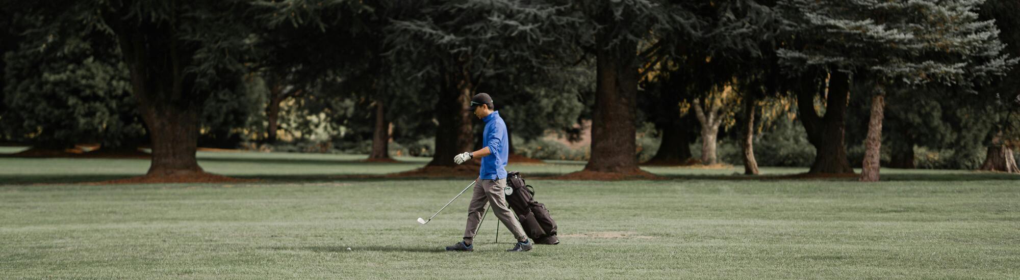 A person wearing a blue shirt and cap walks across a grassy golf course, pulling a golf bag on wheels, with large trees and greenery in the background.