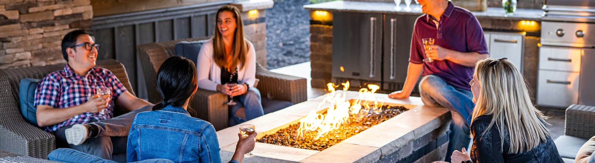 Four adults sit around a rectangular outdoor fire pit, holding drinks and smiling. The setting includes wicker furniture and an outdoor kitchen with a grill in the background.