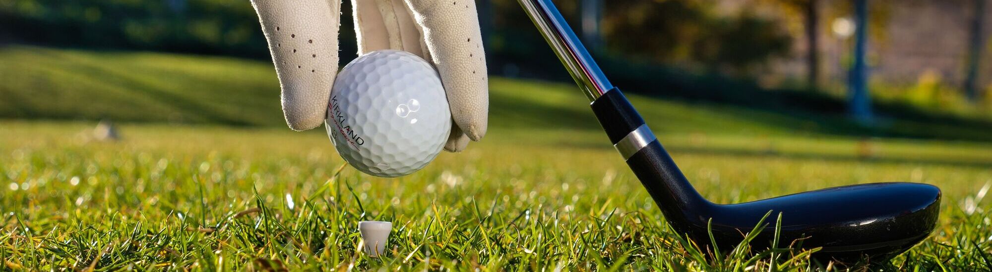 A close-up of a gloved hand placing a golf ball on a tee in the grass, with a golf club positioned nearby, ready for a shot on a sunny day.