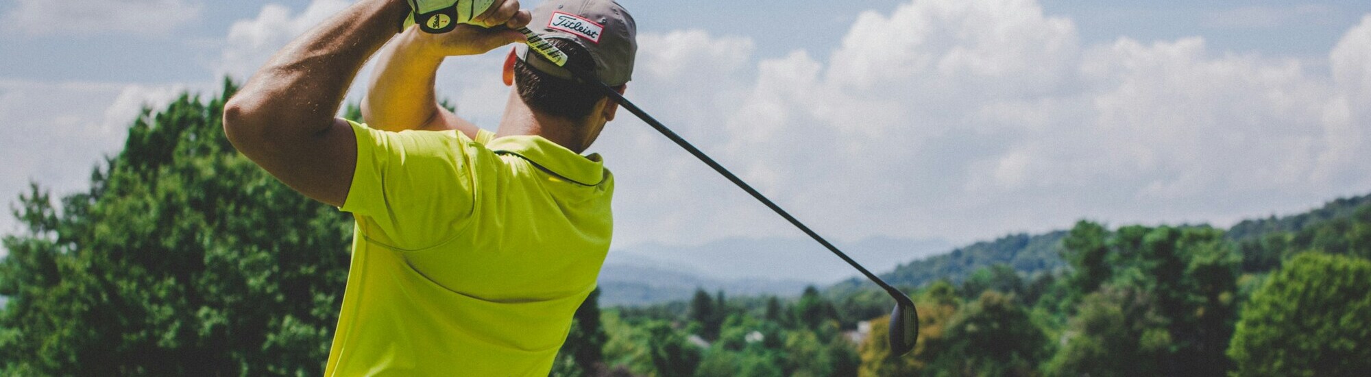A golfer wearing a yellow shirt and cap swings a golf club on a lush, green course with trees and hills in the background under a partly cloudy sky.