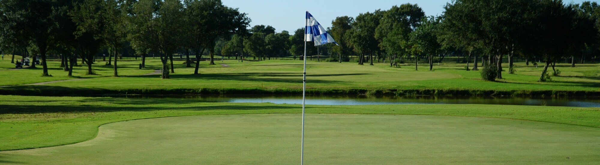 A golf course with a blue and white checkered flag on the green, surrounded by trees and a narrow water hazard in the background under a clear sky.