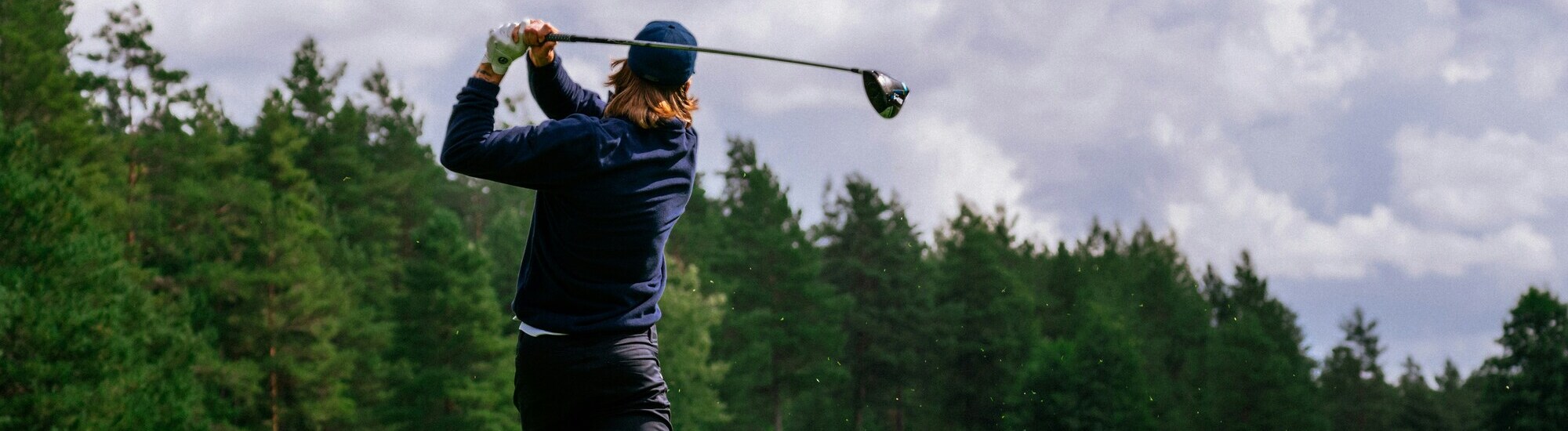 A person wearing a dark jacket and cap swings a golf club on a lush, green golf course surrounded by trees under a cloudy sky.