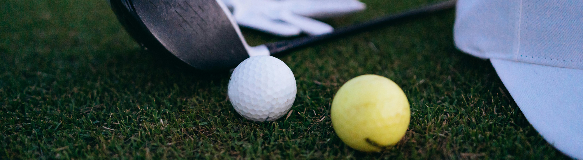 Close-up of a white golf ball and a yellow golf ball on grass, with a golf club, a white glove, and part of a white cap visible in the background.
