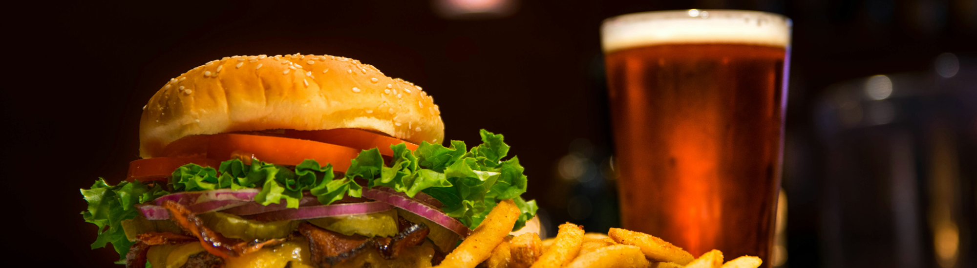 A cheeseburger with lettuce, tomato, onions, and pickles is served with a side of French fries. A glass of amber beer is in the background, all set against a dark blurred backdrop.