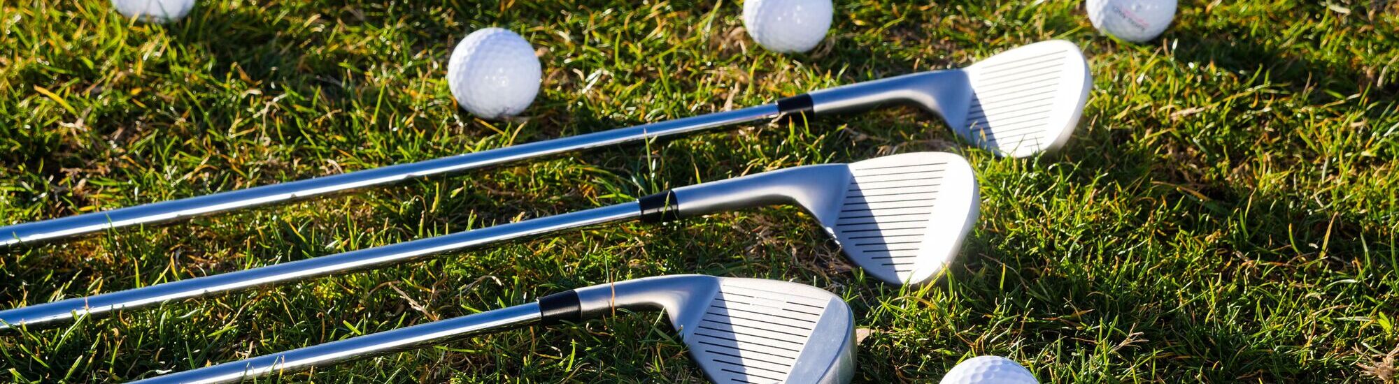Three golf clubs are lying on green grass next to several scattered white golf balls under sunlight.